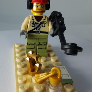 May include: A Lego minifigure of a treasure hunter wearing a red hard hat, headphones, and glasses, holding a metal detector. Gold-colored crystals and a key are on a tan base. The figure is dressed in a tan shirt and green pants.