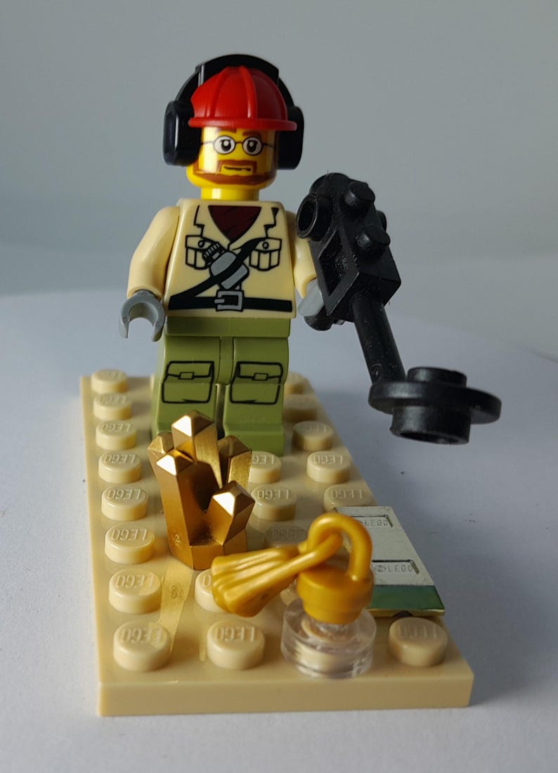 May include: A Lego minifigure of a treasure hunter wearing a red hard hat, headphones, and glasses, holding a metal detector. Gold-colored crystals and a key are on a tan base. The figure is dressed in a tan shirt and green pants.