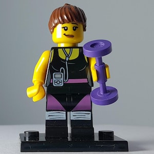 Puede incluir: Una minifigura de Lego con cabello castaño y cabeza amarilla, que viste una camiseta sin mangas negra con un reproductor de música impreso y auriculares blancos. La figura tiene pantalones cortos negros sobre mallas moradas, calentadores blancos y sostiene una mancuerna morada.