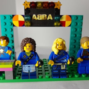 Puede incluir: Un set de construcción LEGO con cuatro minifiguras sobre un escenario de placa base verde. El fondo muestra el texto ABBA en letras doradas con coloridos acentos circulares y de estrellas. Las figuras incluyen un teclista, dos cantantes con micrófonos y un guitarrista con un instrumento azul claro, todos vestidos con trajes azules.