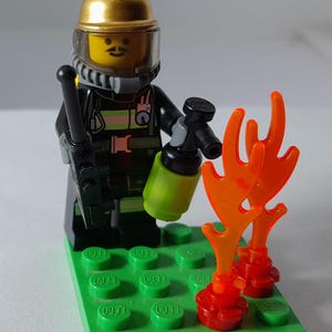 May include: A LEGO firefighter minifigure wearing a gold helmet and black uniform with reflective stripes. The figure holds a black radio and a lime green fire extinguisher. Two translucent orange flame pieces are attached to a green base plate.