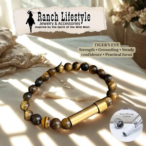 Może przedstawiać: Bransoletka z koralików z tygrysiego oka ze złotym, cylindrycznym zapięciem. Bransoletka jest częścią kolekcji Ranch Lifestyle Jewelry & Accessories, inspirowanej duchem Dzikiego Zachodu. Obraz zawiera również tekst "TIGER'S EYE" i "Secret Message Cylinder."