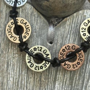 May include: Close-up of a bracelet featuring circular metal beads with the text "GAUGE" and "12 G" or "2 G" in a stamped font. The beads are connected by a black leather cord. The beads are in gold and copper tones.