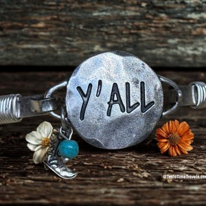 May include: A silver bracelet with the word "Y'ALL" engraved on a circular charm. The bracelet features a turquoise bead, a small silver boot charm, and a white flower. An orange flower is also present.