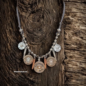 Puede incluir: Un collar artesanal con colgantes Winchester 45 Auto y 12 Gauge Shotgun Shell. El collar tiene un cordón gris y detalles metálicos plateados. Los colgantes están adornados con pequeñas piedras brillantes.