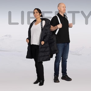 Op de afbeelding: Twee mensen staan voor een besneeuwde bergachtergrond. De vrouw draagt een zwarte pufferjas en de man draagt een zwart poloshirt en jeans. Het woord "LIBERTY" staat in grote letters achter hen.