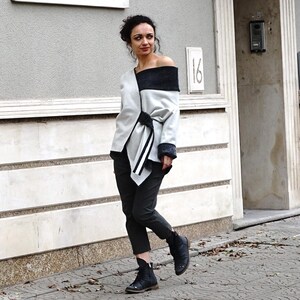 May include: A woman wearing a light gray off-the-shoulder jacket with a black collar and a black belt. The jacket has a white panel with black and white stripes. She is wearing dark gray pants and black boots.