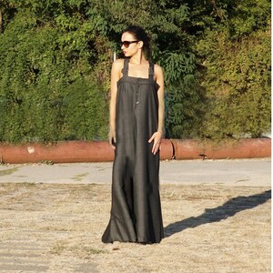 May include: A dark gray maxi dress with wide straps and large buttons down the front. The dress has a loose, flowing silhouette and is made of a textured fabric. The model is wearing sunglasses and standing outside in front of green foliage.