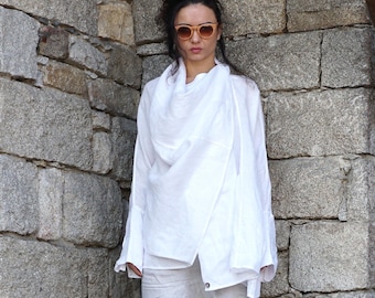 May include: A woman wearing a white linen wrap top and matching pants. The top has a draped neckline and long sleeves. She is also wearing light brown sunglasses. The background is a stone wall.