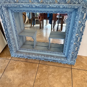 Può includere: Uno specchio decorativo, ornato, con cornice azzurra e due piccoli ripiani all'interno. La cornice presenta intagli dettagliati e una finitura anticata. Lo specchio riflette una stanza con mobili. È un elemento di arredo murale.