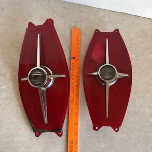 May include: Two vintage car tail light lenses. The lenses are a deep red colour with a chrome emblem in the centre. The emblem has a circular centre with a cross-like design extending outwards. A ruler is placed between the lenses.