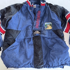Puede incluir: Chaqueta anorak vintage azul y negra con la palabra "PANTHERS" bordada en rojo. La chaqueta tiene un bolsillo delantero, un pequeño parche con una escena de montaña y un logotipo de tigre. Las mangas tienen rayas rojas y blancas.