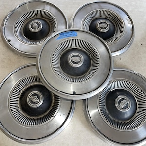 Puede incluir: Cinco tapacubos plateados vintage con un centro negro y un logotipo. Los tapacubos tienen un diseño acanalado alrededor del centro y una superficie brillante y reflectante. Los tapacubos son para una rueda de 35,56 cm.