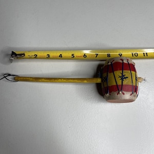 May include: A small, hand-held drum with a yellow handle. The drum is decorated with red, yellow, and blue stripes and a star design. The drum is approximately 4 inches in diameter.