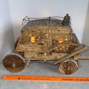 Op de afbeelding: Een rustiek houten Wells Fargo postkoetsmodel met het jaartal 1854 op de zijkant. Het model heeft een verweerde uitstraling, met zichtbare houtnerf en een metalen dak. De wielen zijn van hout. Het model is ongeveer 30 cm lang.
