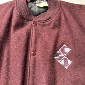 May include: A maroon wool varsity jacket with a ribbed collar and cuffs. The jacket features a button-up front and a diamond-shaped logo on the chest. The jacket has a quilted lining and a tag that says "BUTWIN".