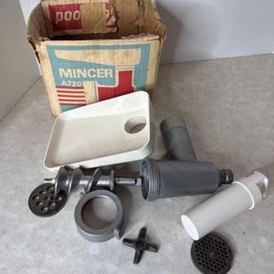 May include: A disassembled vintage meat mincer with various parts, including a white plastic tray, a metal auger, and grinding plates. The original cardboard box is visible, with the word "MINCER" and model number "A720" printed on it.