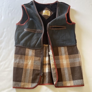 May include: A vintage vest with a dark gray upper section and a brown and tan plaid lower section. The vest has a brown leather trim and a dark brown, fuzzy interior. A small leather patch with text is visible on the chest.