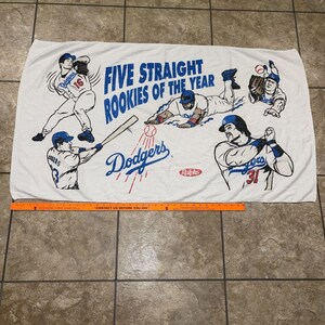 Peut inclure: Serviette de plage blanche avec un logo des Dodgers de Los Angeles en bleu et rouge et le texte "Five Straight Rookies of the Year". La serviette présente des illustrations de cinq joueurs de baseball portant des uniformes des Dodgers.