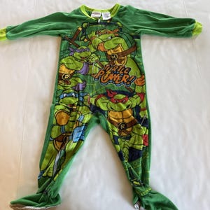 May include: Green one-piece pajama featuring the Teenage Mutant Ninja Turtles. The pajama has long sleeves and attached feet. The front of the pajama displays the phrase "Turtle Power!" and images of the four turtles.