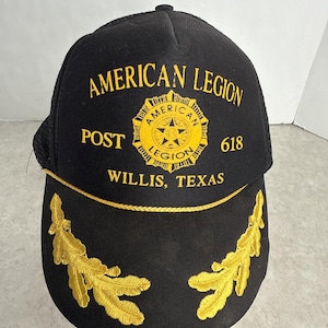 May include: Black baseball cap with the words "AMERICAN LEGION POST 618 WILLIS, TEXAS" in gold lettering. The cap features a gold emblem and gold leaf embroidery on the brim. The back of the cap is black mesh.