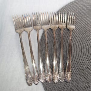 May include: A set of seven vintage silver forks with tines and ornate handles. The forks are arranged on a gray and white surface, showcasing their aged patina and detailed design. Ideal for antique silverware collectors.