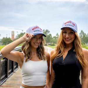 Puede incluir: Dos gorras de béisbol con el texto "St. Louis Show Me State" en rojo y azul. Las gorras tienen un diseño de rayas blancas y azules. Las gorras las llevan dos personas en un puente de madera.