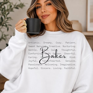 May include: A white long-sleeve sweatshirt featuring black text that reads: Artistic. Dreamy. Cozy. Patient. Sweet Natured. Creative. Nurturing. Gentle. Joyful. Calm. Thoughtful. Comforting. Soft Spoken. Cheerful. Caring. Honest. Wholesome. Kind. Baker.