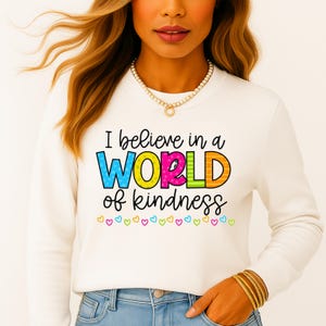 Puede incluir: Una sudadera blanca con el texto "I believe in a WORLD of kindness" en letras coloridas. La palabra "WORLD" está en azul, amarillo, rosa, naranja y verde. Pequeños corazones de varios colores en la parte inferior.