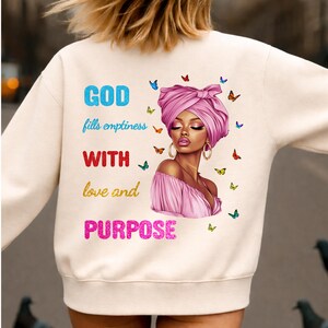 Pode incluir: Camisola creme com o texto "GOD fills emptiness WITH love and PURPOSE" em azul, vermelho e rosa. Um gráfico de uma mulher com um turbante e vestido rosa está à direita, rodeada por borboletas.