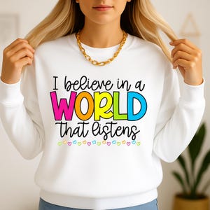 Puede incluir: Sudadera blanca con la frase "I believe in a WORLD that listens" en letras coloridas. La palabra "WORLD" es rosa, naranja, amarilla y azul. Pequeños corazones coloridos en la parte inferior del diseño.