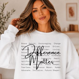 Può includere: Felpa girocollo bianca con le parole "Difference Matter" in un carattere scritto a mano. La felpa presenta anche un elenco di parole positive come "Brave Spirit", "Encouraging" e "Kindness".