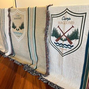 May include: Several beige and cream-colored camp blankets with decorative fringe. Each blanket features a shield design with the camp name and imagery of trees, oars, and water. The blankets have vertical stripes in green, blue, and burgundy.
