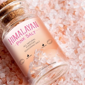 Peut inclure: Un pot en verre de sel rose de l'Himalaya. L'étiquette indique "Sel rose de l'Himalaya" et "Sel d'assaisonnement quotidien entièrement naturel". Le pot est posé sur un lit de cristaux de sel rose de l'Himalaya.
