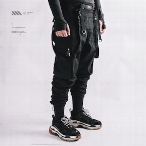 May include: Black cargo pants with multiple straps and pockets. The pants have a high waist and are paired with black sneakers with white and red accents. The outfit is completed with a long-sleeved black shirt.