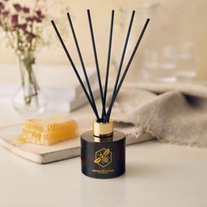 Op de afbeelding: Een bruine glazen diffuserfles met zwarte rietjes en een gouden dop. De fles heeft een gouden etiket met een bij en honingraatontwerp en de tekst "The Beekeeper Collection".