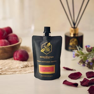 Puede incluir: Una bolsa negra de recarga de difusor de caña "The Beekeeper Collection", etiquetada como "Wild Orchard". La bolsa tiene un logotipo de abeja dorada y una tapa negra. Un frasco difusor de vidrio con cañas y un cuenco de ciruelas rojas están en el fondo.