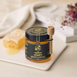May include: A small glass jar of honey with a black label that reads "The Beekeeper Collection Honey Limited Edition Raw Natural Honey Made by Bees in Shropshire, England 275g". The jar is on a white surface with a wooden honey dipper.