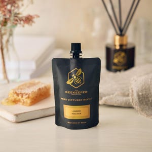 Puede incluir: Una bolsa negra de recarga para difusor de varillas, con texto dorado y un logotipo de abeja. Dice "Amber Nectar" e "Inspired by Bees". Un panal y un difusor de varillas están en el fondo. De la colección The Beekeeper.