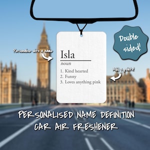 May include: A white car air freshener with the name "Isla" and a definition of the name. The definition says Isla is kind hearted, funny, and loves anything pink. The air freshener is hanging from a rearview mirror in a car.