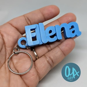 May include: A light blue keychain spelling "Elena" in 3D block letters. The keychain includes a silver metal split ring. The keychain is held in a hand. A teal circle logo with the text "OA Gift" is in the bottom right corner.