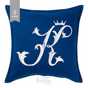 Pode incluir: Uma almofada decorativa azul royal com a letra "K" bordada em branco, encimada por uma coroa. A letra está em uma fonte cursiva ornamentada. A almofada é quadrada e tem uma textura macia. O texto "6 tamanhos" também é visível.