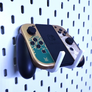 Könnte beinhalten: Eine Nintendo Switch-Konsole mit goldfarbenen und türkisfarbenen Joy-Cons ist an einer weißen Lochwand befestigt. Die Joy-Cons haben dekorative Muster und schwarze Knöpfe. Ein schwarzer Griff ist am linken Joy-Con befestigt.