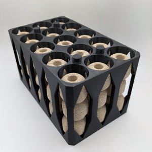 May include: A black container with a geometric design, filled with beige, ring-shaped objects. The container has a rectangular base and diamond-shaped cutouts on the sides. The objects are arranged in circular openings.