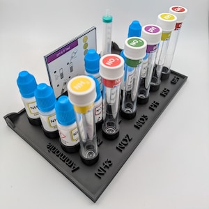 May include: A black plastic test tube rack holding several clear glass tubes and small bottles with blue, red, green, yellow, and white caps. The rack also holds a test card with instructions. The bottles and tubes are labelled with chemical symbols.