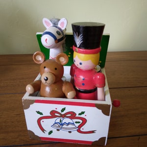 May include: A white wooden music box with a red, green, and white Christmas design. The box has a crank on the side and features a teddy bear, a horse, and a toy soldier inside.