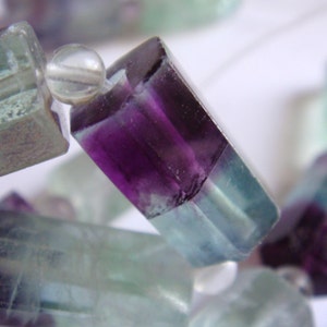 May include: A close-up of a strand of faceted fluorite beads. The beads are a mix of purple, green, and clear colors. The beads are strung on a clear thread.