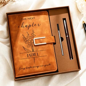 May include: A tan leather-effect journal in a gift box featuring the text THE NEXT chapter, a floral illustration, the name JADIEL, and the phrase Every chapter brings new growth. The set includes a matching pen and a refill ink cartridge.