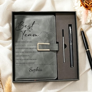 Op de afbeelding: Een grijs notitieboek met een goudkleurige metalen sluiting, voorzien van de tekst Best Team, een hartomtrek en de naam Sophia. De set bevat een bijpassende grijze pen en een vulling, gepresenteerd in een zwarte geschenkdoos met inzetstuk.