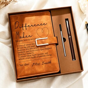 May include: A tan notebook with a metal clasp and the text Difference Maker, a heart outline, and a definition printed on the cover. The notebook is inside a brown gift box with a matching pen and a refill cartridge, plus an extra black pen.